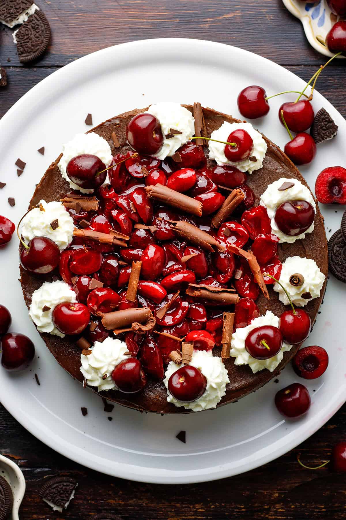 Black Forest Cheesecake A chocolate cheesecake topped with whipped cream, fresh cherries, cherry glaze, and chocolate shavings sits on a white plate.
