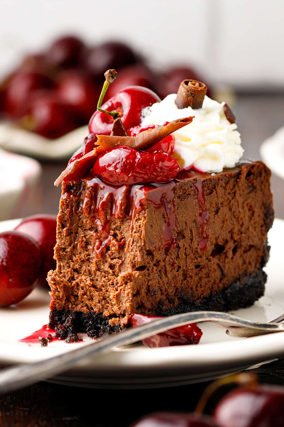 Black Forest Cheesecake A slice of chocolate cheesecake topped with cherry sauce, whipped cream, a cherry, and chocolate curls, served on a white plate.