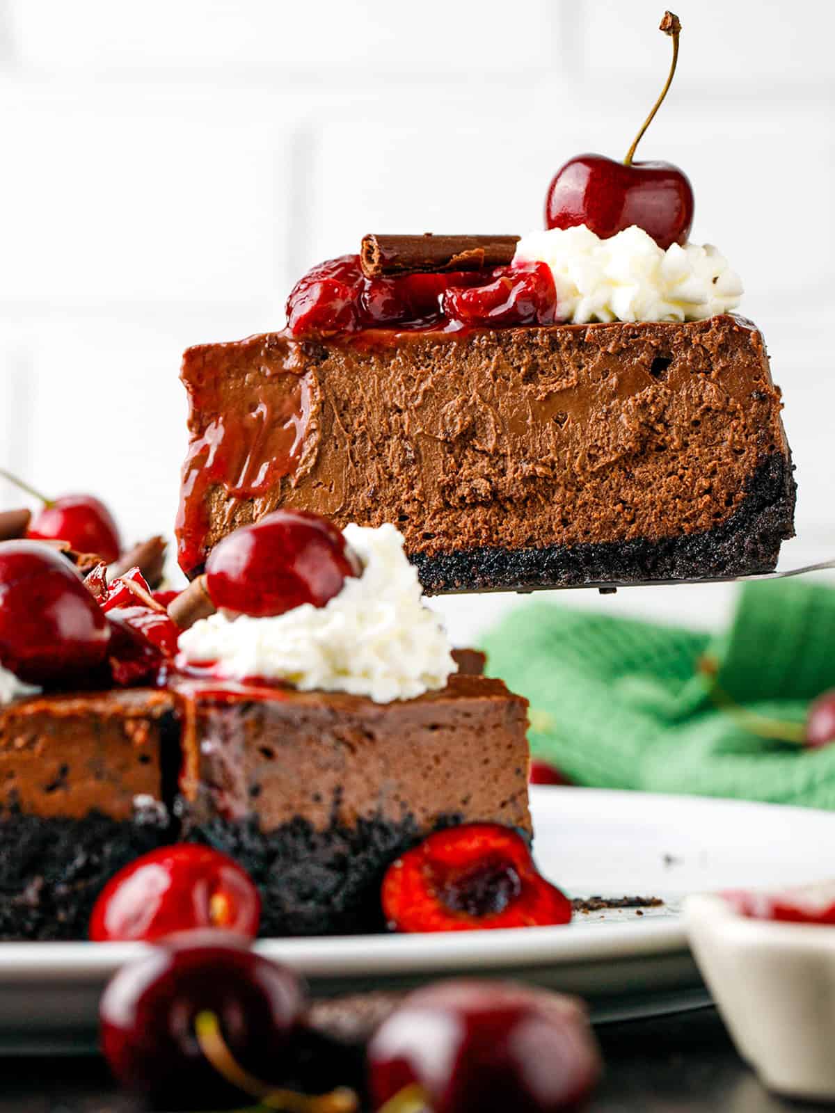 Black Forest Cheesecake A slice of rich chocolate cheesecake with whipped cream, cherries, and a chocolate garnish, being lifted from a whole cheesecake with a dark cookie crust.