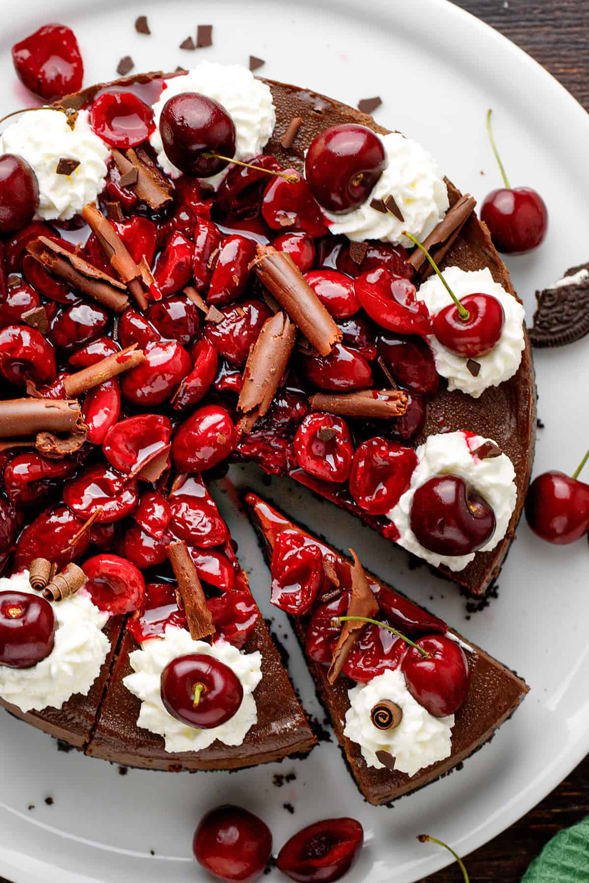 Black Forest Cheesecake A chocolate cheesecake topped with cherries, dollops of whipped cream, and chocolate shavings sits on a white plate. One slice is slightly pulled out.