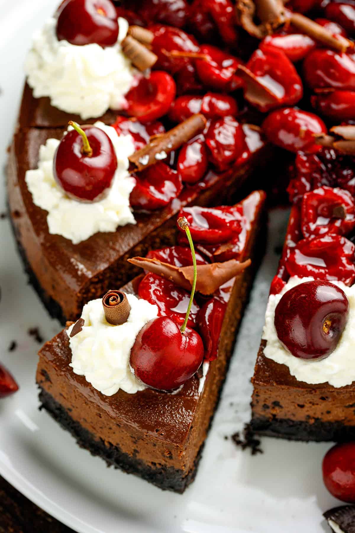 Black Forest Cheesecake A chocolate cheesecake topped with whipped cream, fresh cherries, chocolate curls, and glossy cherry sauce, with one slice slightly pulled out from the cake.