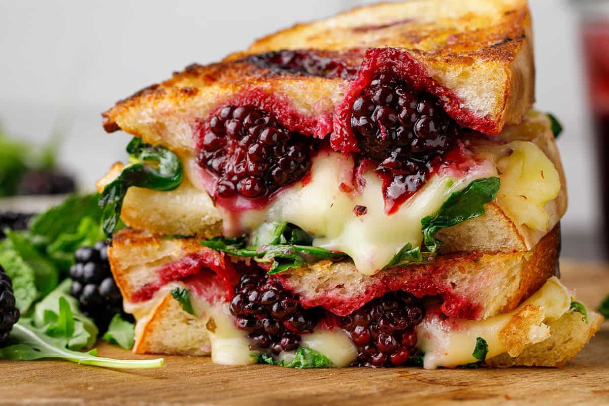 A close-up of a grilled cheese sandwich filled with melted cheese, fresh blackberries, and greens.