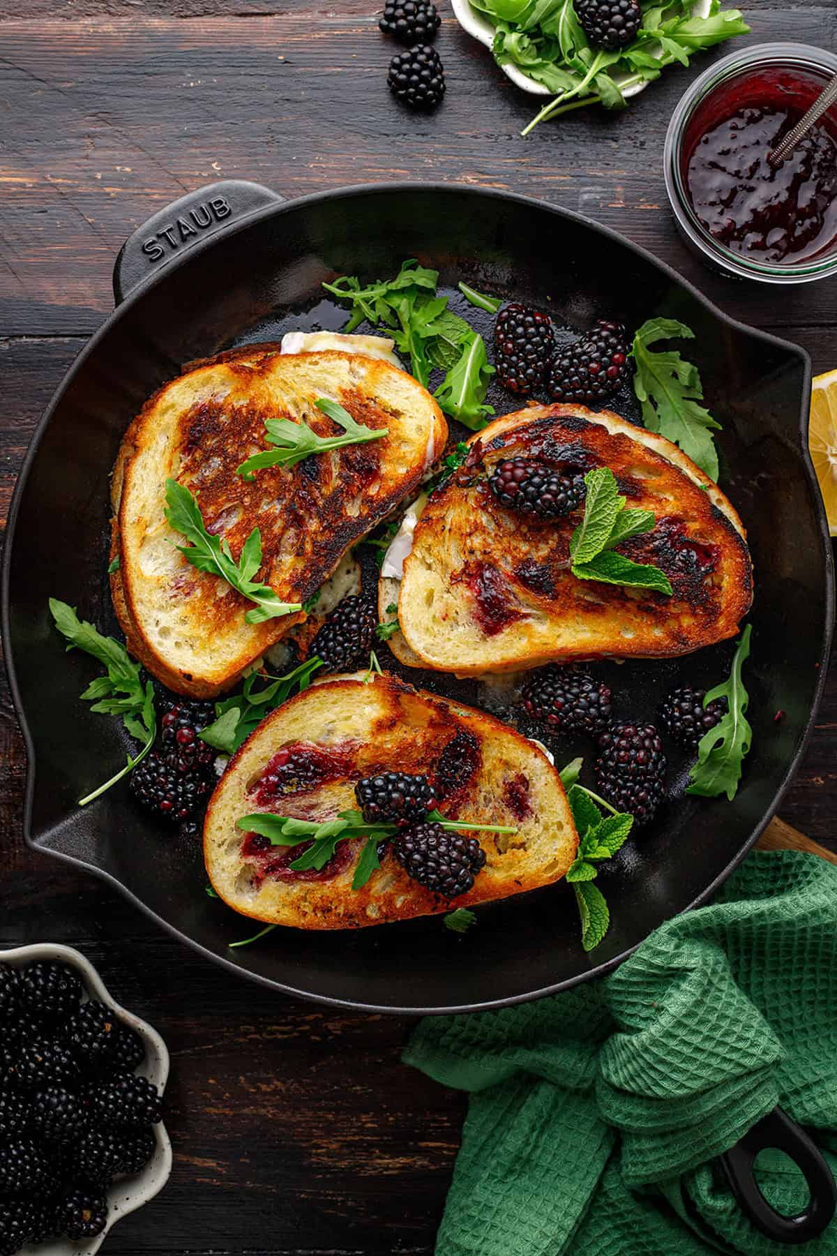 Three slices of toasted bread with blackberry jam, fresh blackberries, arugula, and mint in a black skillet.