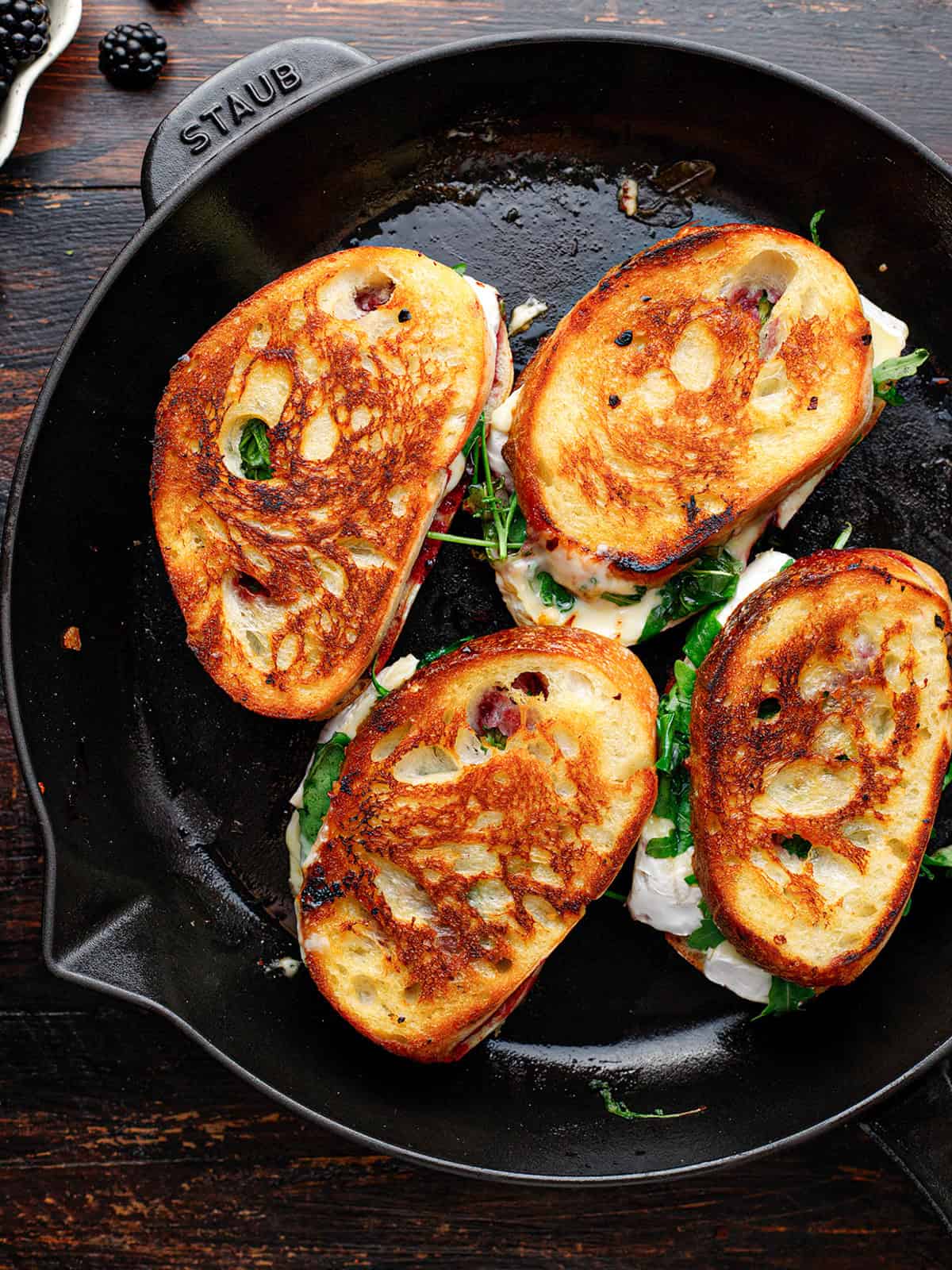 Four grilled sandwiches with golden bread and leafy greens are cooking.