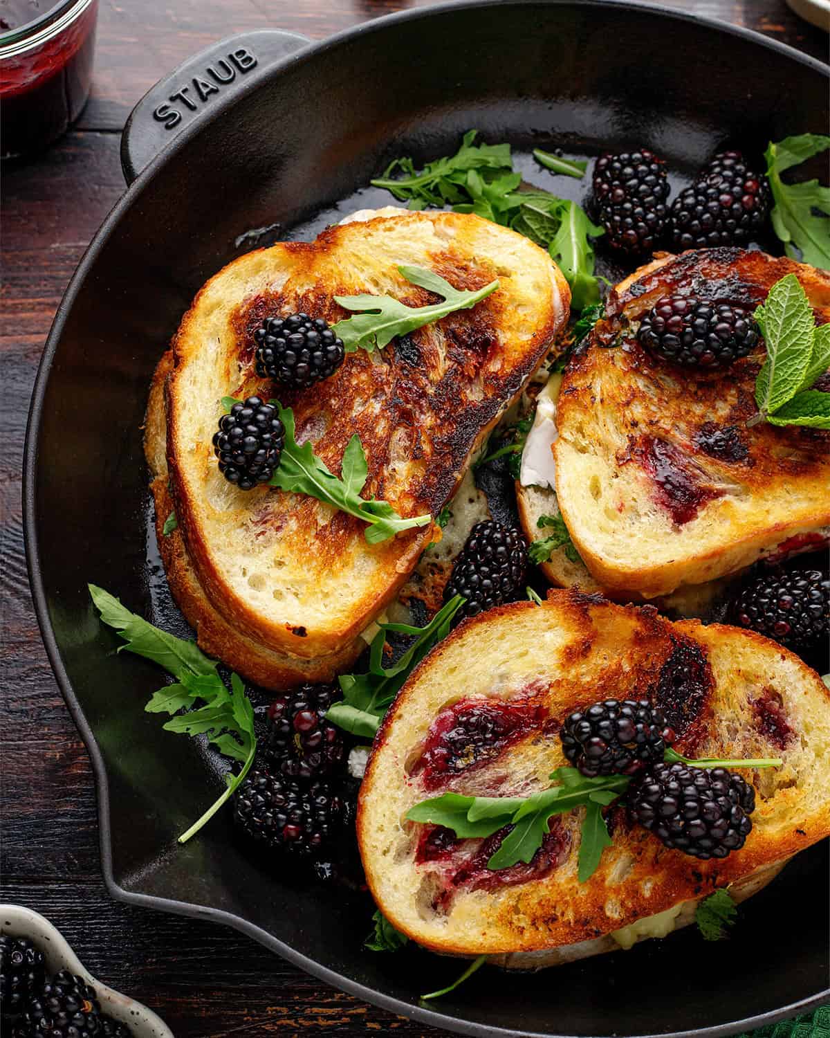 Three grilled cheese sandwiches with blackberry jam, fresh blackberries, arugula, and mint leaves are arranged in a cast iron skillet.