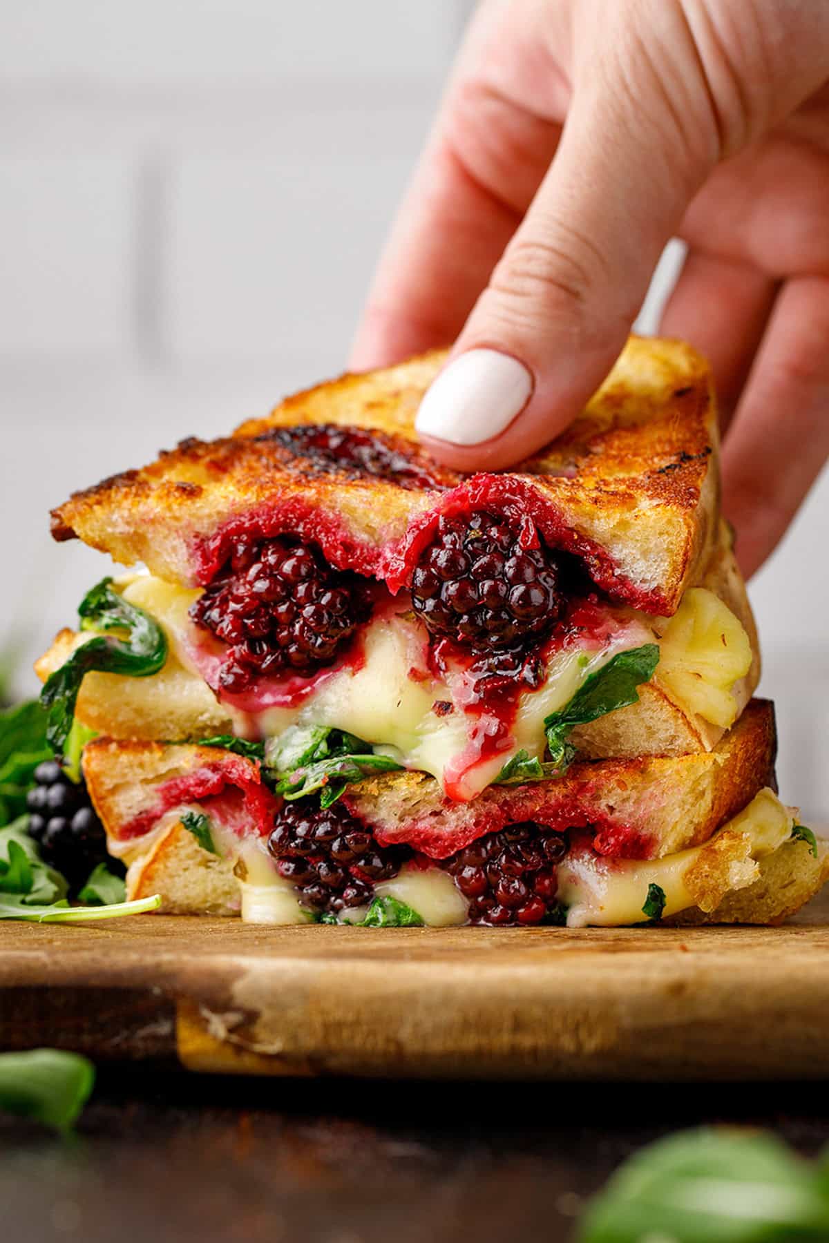 A hand holds a stacked grilled cheese sandwich with melted cheese, fresh blackberries, and greens.
