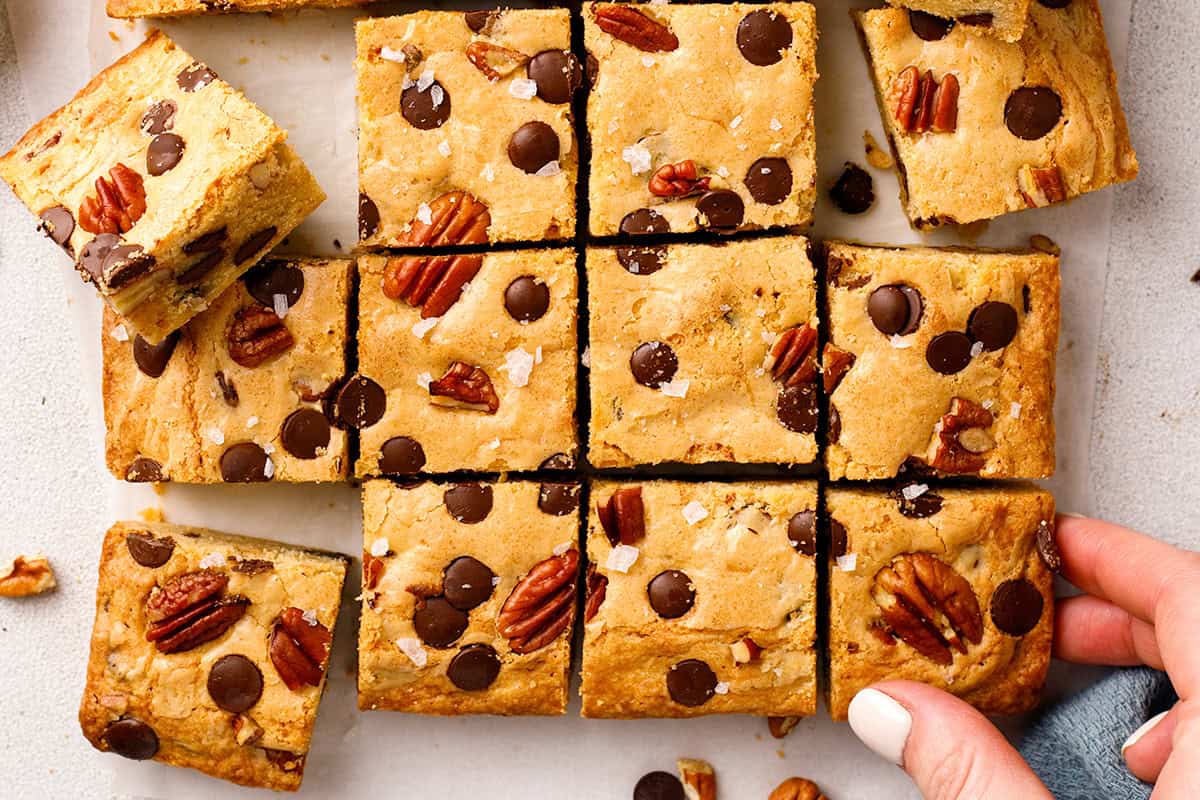 Blondies Golden brown chocolate chip cookie bars (blondies) topped with pecans are cut into squares on a white surface.