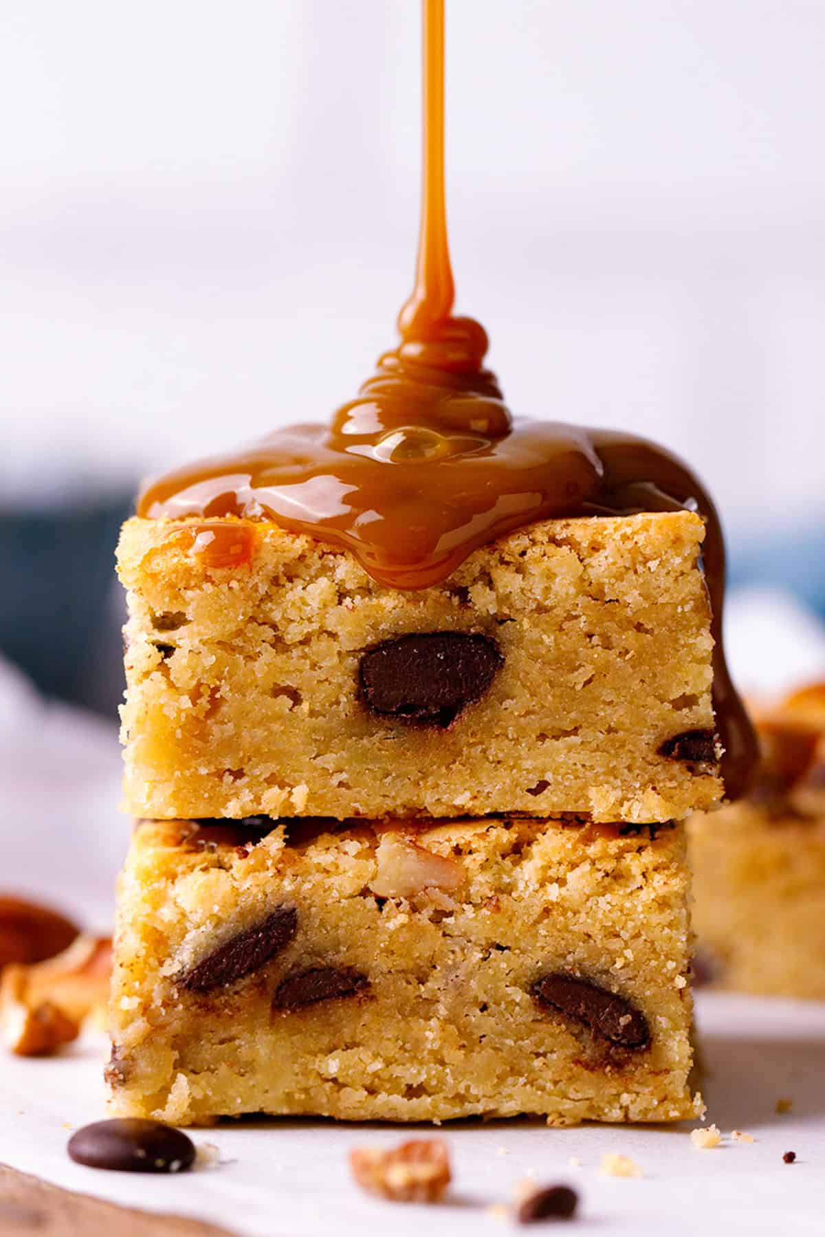 Blondies Two thick blondie squares with chocolate chips are stacked, topped with a generous drizzle of caramel sauce pouring down the sides.