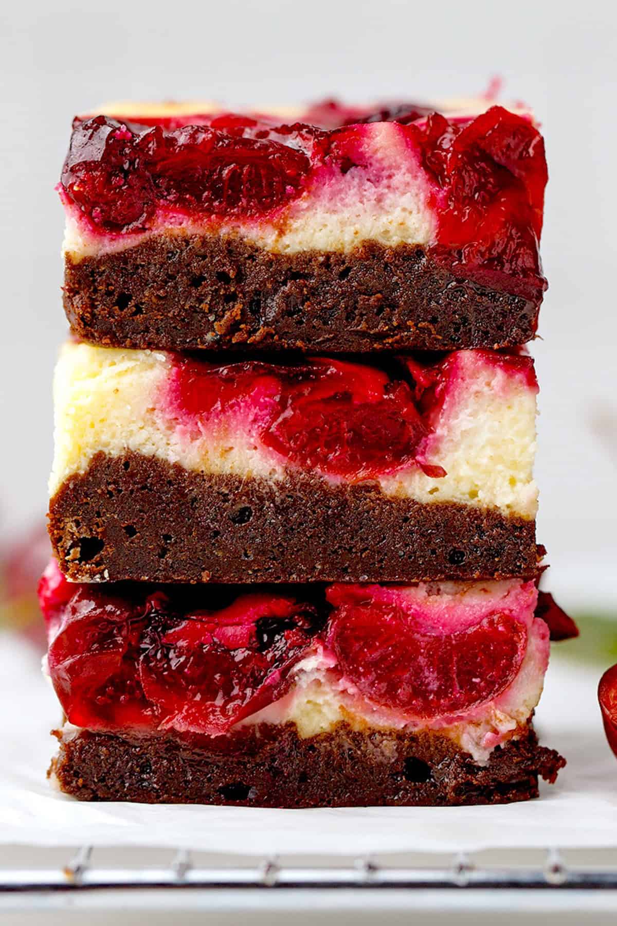 Cherry Cheesecake Brownies Three stacked dessert bars with a brownie base, creamy cheesecake layer, and a glossy cherry topping, shown close-up with vibrant red cherries visible.