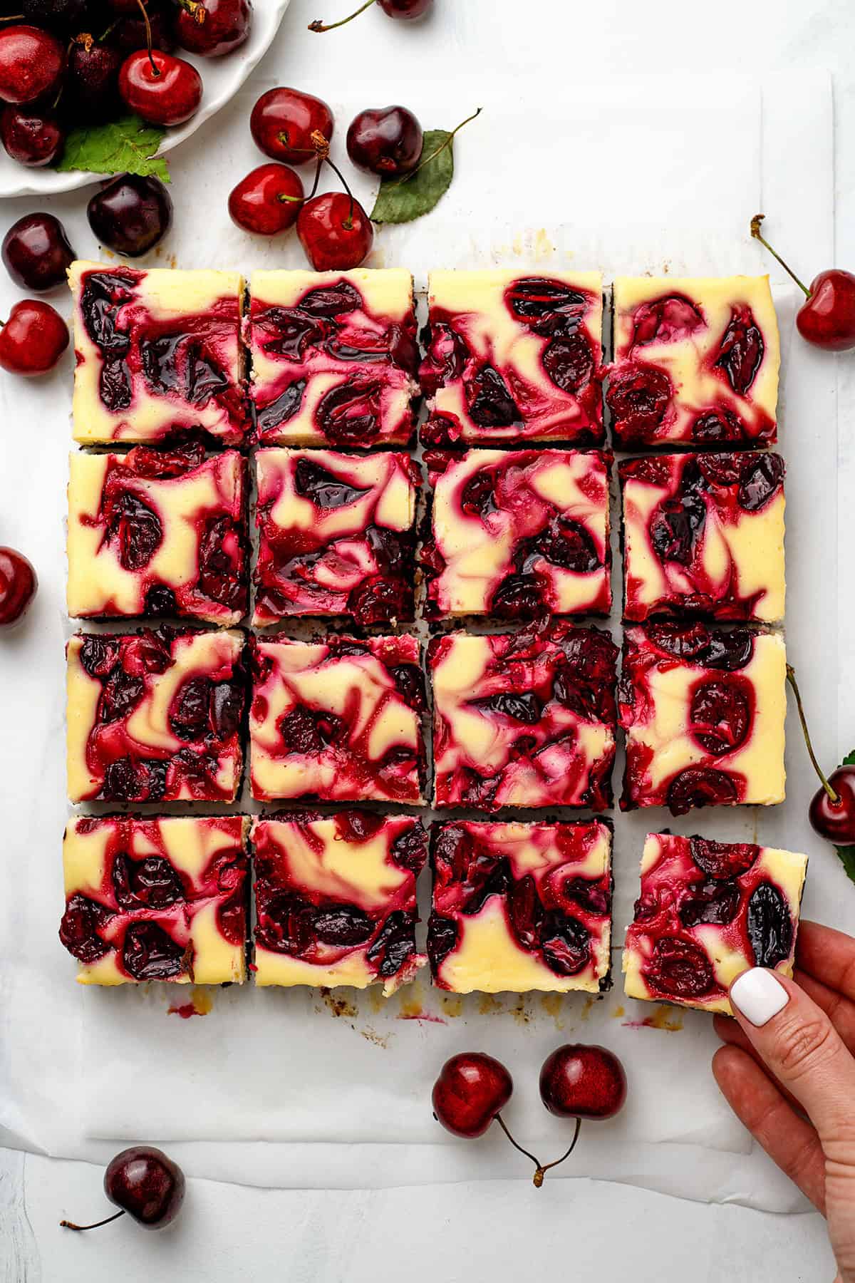Cherry Cheesecake Brownies Cherry swirl cheesecake brownies cut into squares on parchment paper, with one piece being lifted by a hand.