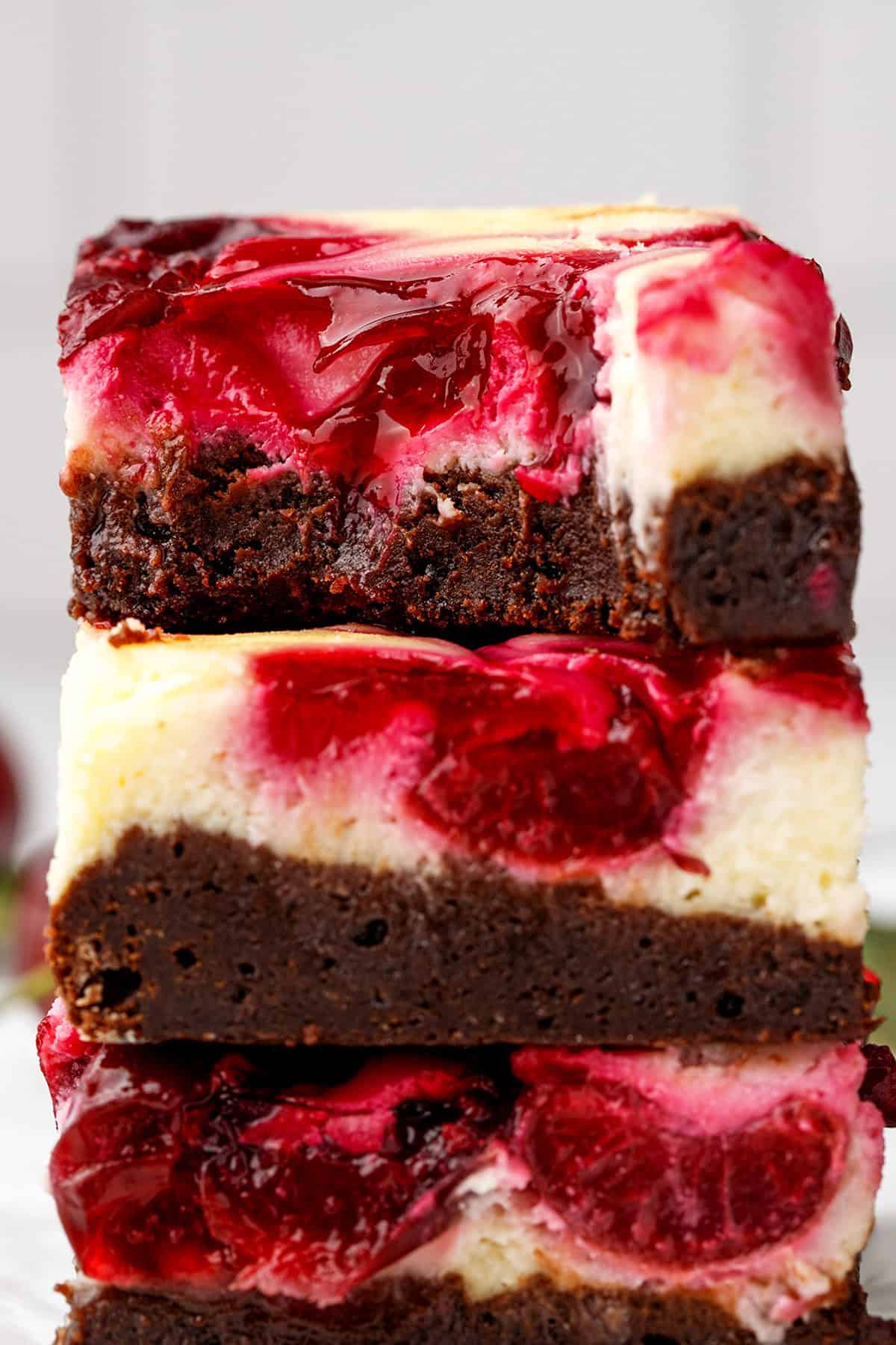 Cherry Cheesecake Brownies Chocolate brownie cheesecake bars stacked, each topped with a vibrant red cherry swirl and creamy cheesecake layer, shot close up to show texture.