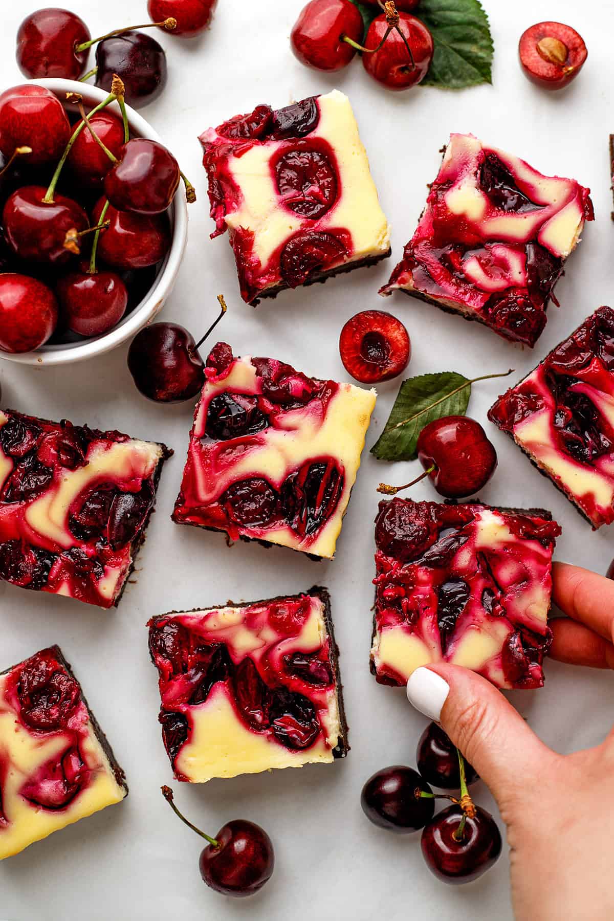 Cherry Cheesecake Brownies Cherry cheesecake brownies with swirled red cherry topping are arranged on a white surface, surrounded by fresh cherries.