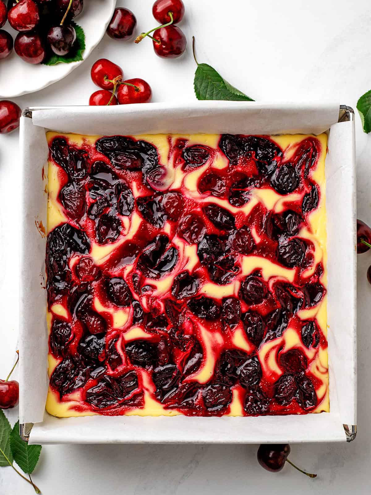 Cherry Cheesecake Brownies A square pan of cherry cheesecake bars with a swirled cherry topping, surrounded by fresh cherries on a white surface.