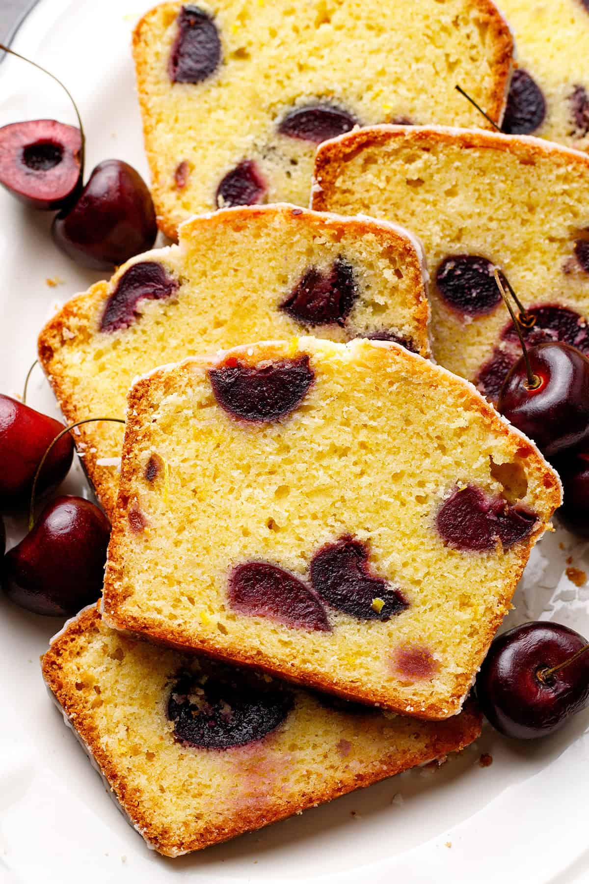 Cherry Loaf Cake Slices of cherry cake with visible cherry pieces are arranged on a white plate. The cake appears moist and golden with a slightly crisp crust.