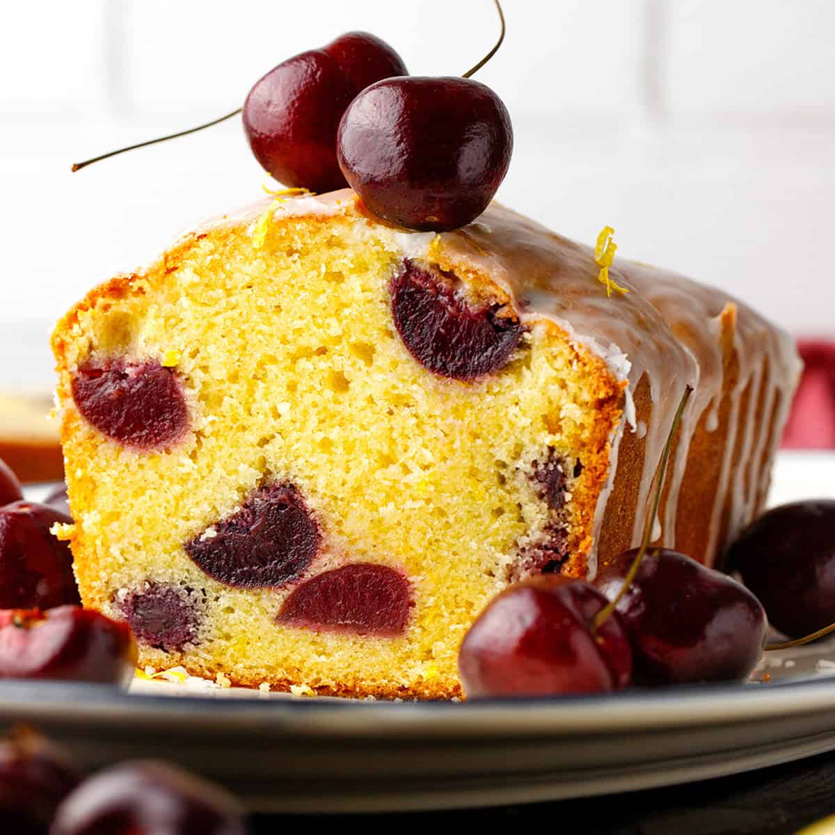 Cherry Loaf Cake A sliced cherry loaf cake with visible cherries inside, topped with icing and fresh cherries, sits on a plate.