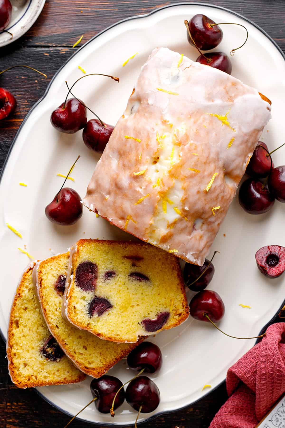 Cherry Loaf Cake A glazed loaf cake with cherries and lemon zest is shown on a white platter. Three slices reveal whole cherries baked inside the golden cake.