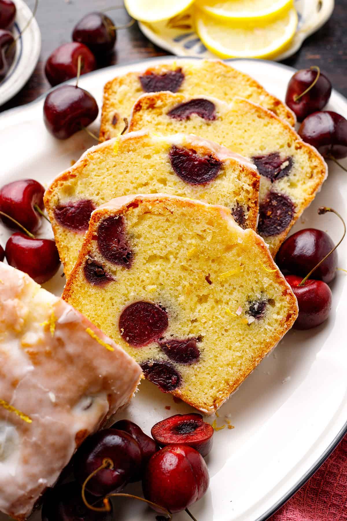 Cherry Loaf Cake Slices of cherry loaf cake are on a white plate, surrounded by fresh cherries and lemon slices.