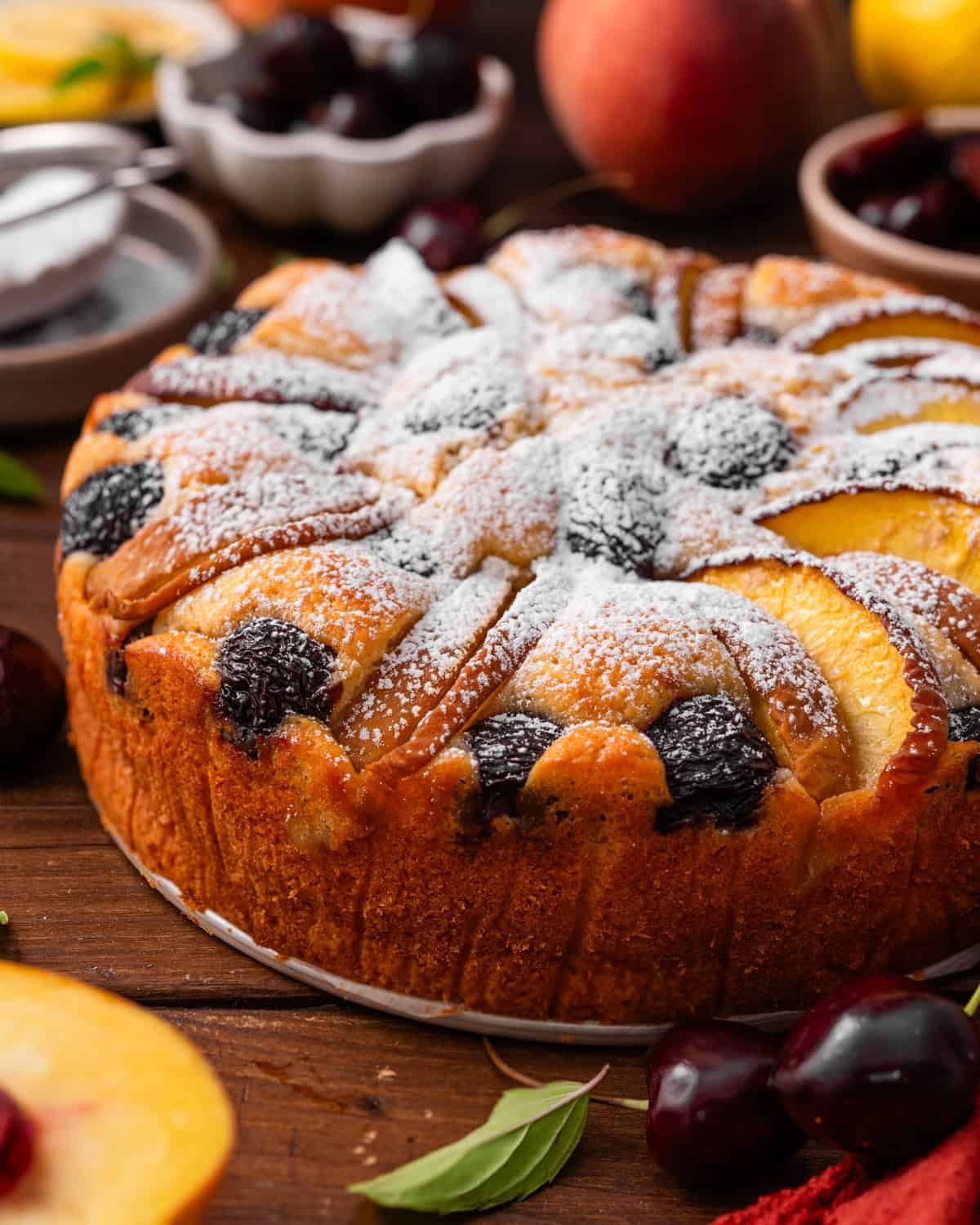Italian-Style Lemon Ricotta Cake with Cherries and Peaches Round fruit cake topped with powdered sugar, peach slices, and cherries.
