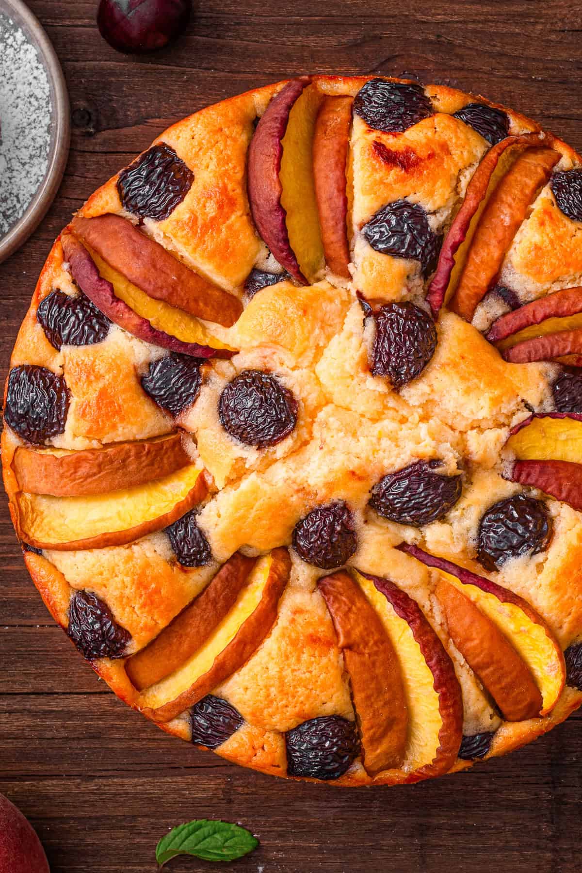 Italian-Style Lemon Ricotta Cake with Cherries and Peaches A round fruit cake topped with alternating slices of peaches and cherries, arranged in a circular pattern, sits on a wooden surface.