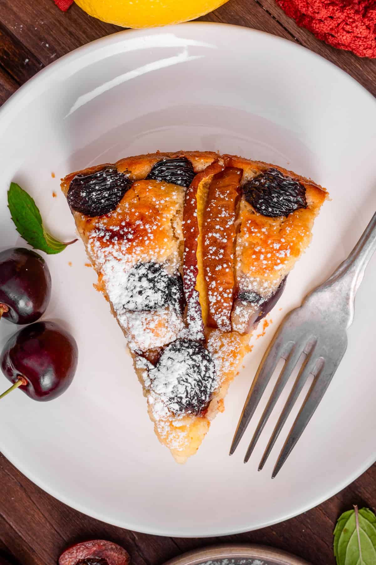 Cherry Peach Ricotta Cake A slice of fruit cake dusted with powdered sugar sits on a white plate with a fork.