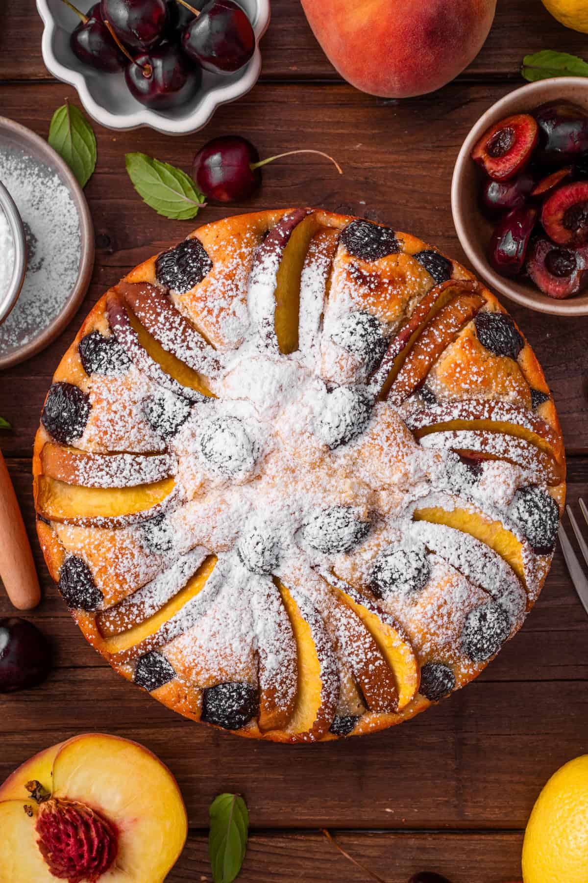 Cherry Peach Ricotta Cake A round cake topped with peach slices and cherries, dusted with powdered sugar.