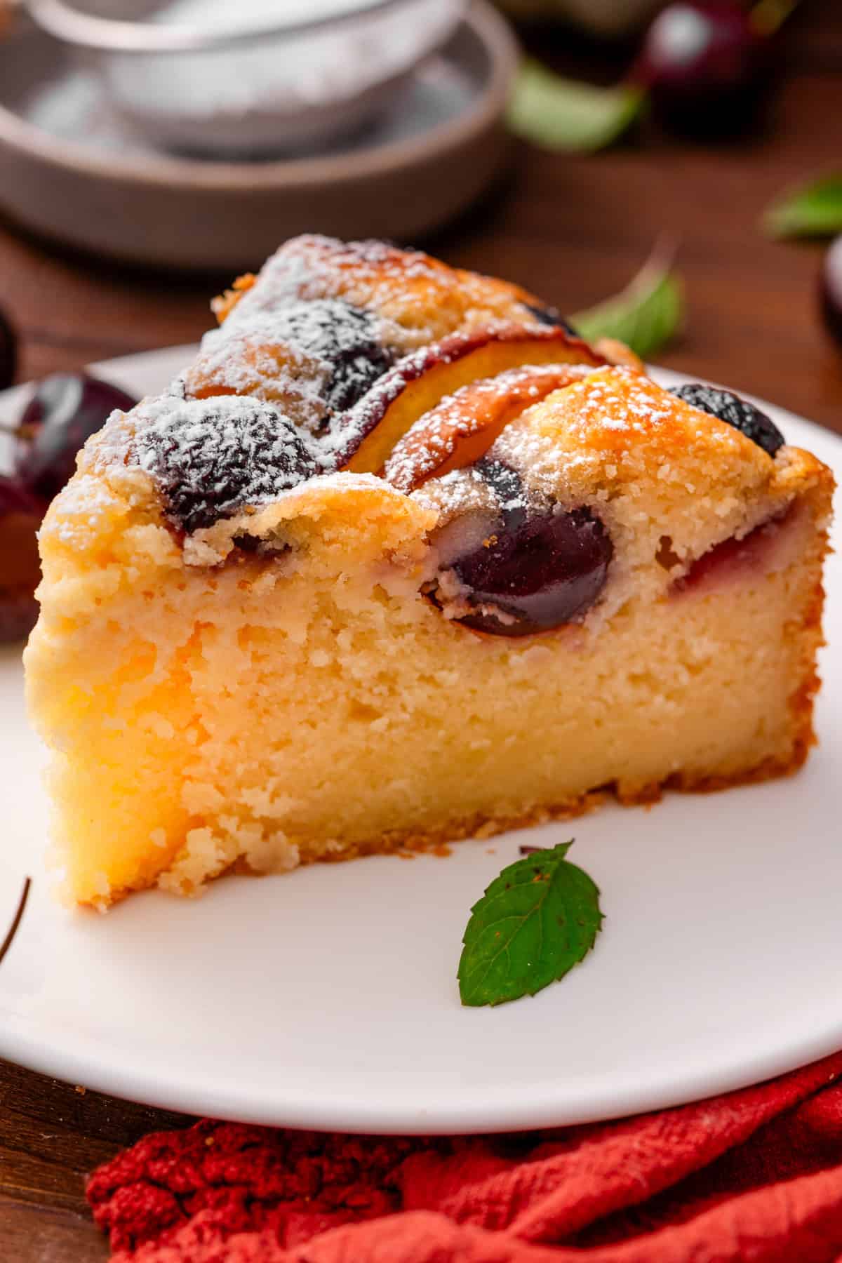 Italian-Style Lemon Ricotta Cake with Cherries and Peaches A close-up of a slice of cherry cake on a white plate, topped with powdered sugar and garnished with a mint leaf.