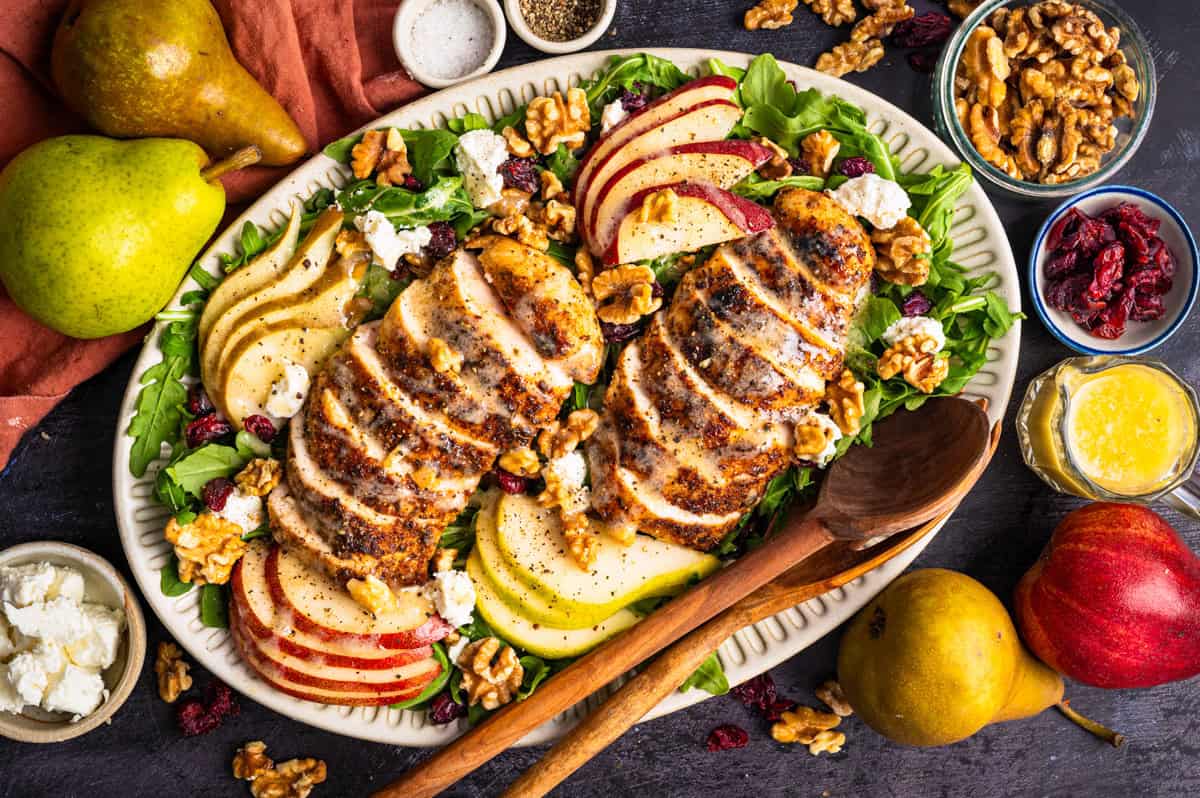 Chicken Arugula Salad with Pears, Goat Cheese, and Walnuts A platter of sliced grilled chicken on greens, garnished with apple and pear slices, walnuts, goat cheese, and dried cranberries, surrounded by ingredients and salad dressing.