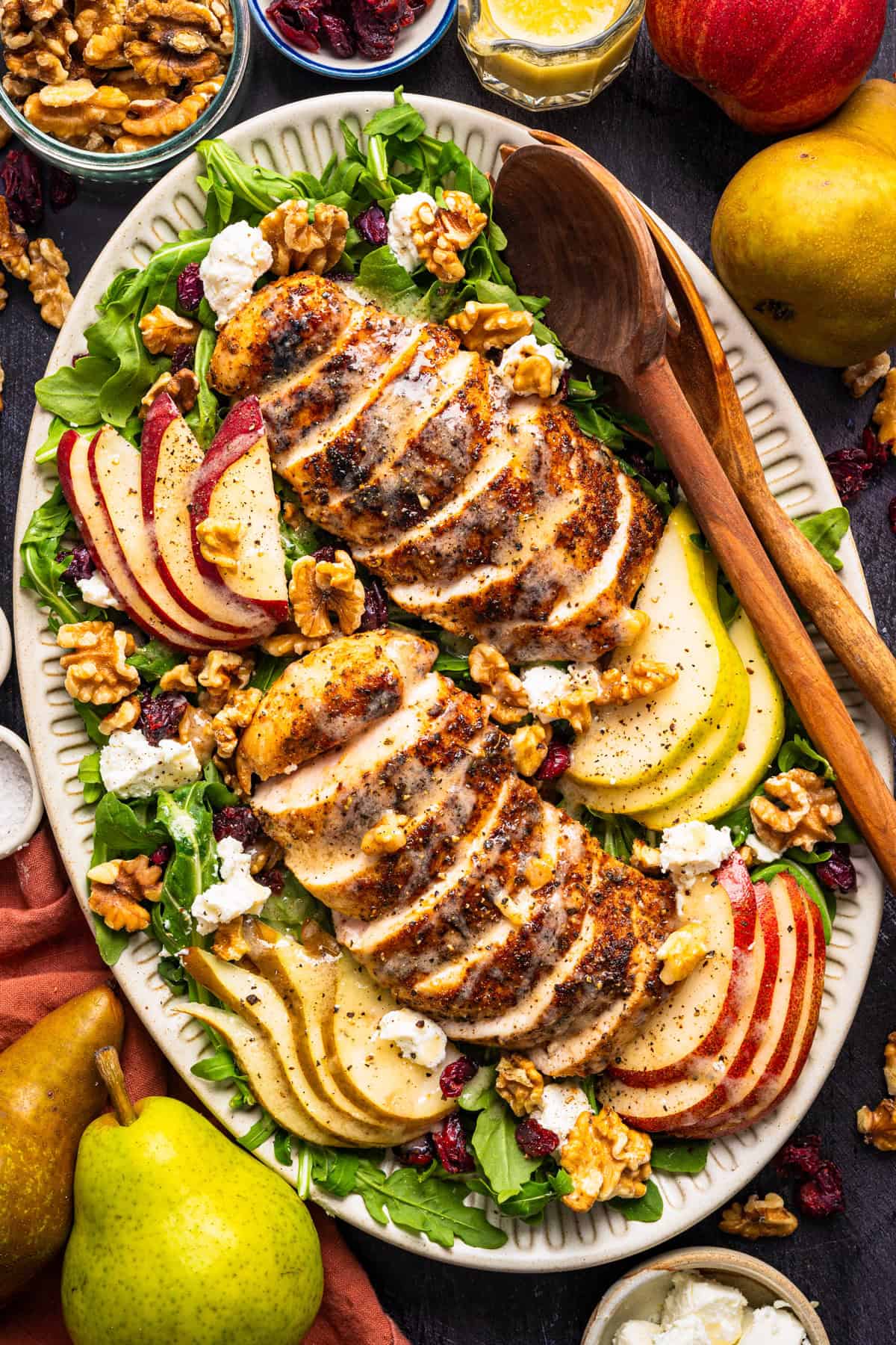 Chicken Arugula Salad Sliced grilled chicken on a bed of arugula, surrounded by apple, pear, walnuts, goat cheese, and dried cranberries on an oval platter.