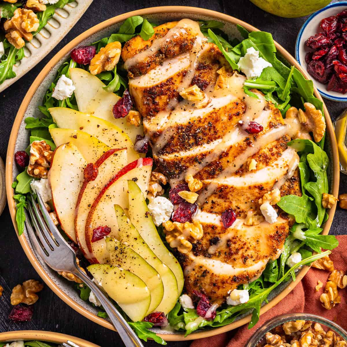 Chicken Arugula Salad with Pears, Goat Cheese, and Walnuts Salad with sliced grilled chicken, fresh pear slices, arugula, walnuts, dried cranberries, and crumbled cheese on a plate.
