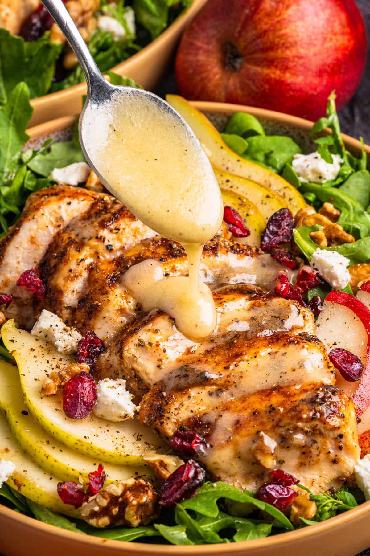 Chicken Arugula Salad A spoon drizzles dressing over a salad with sliced grilled chicken, pear, cranberries, walnuts, goat cheese, and arugula in a bowl.