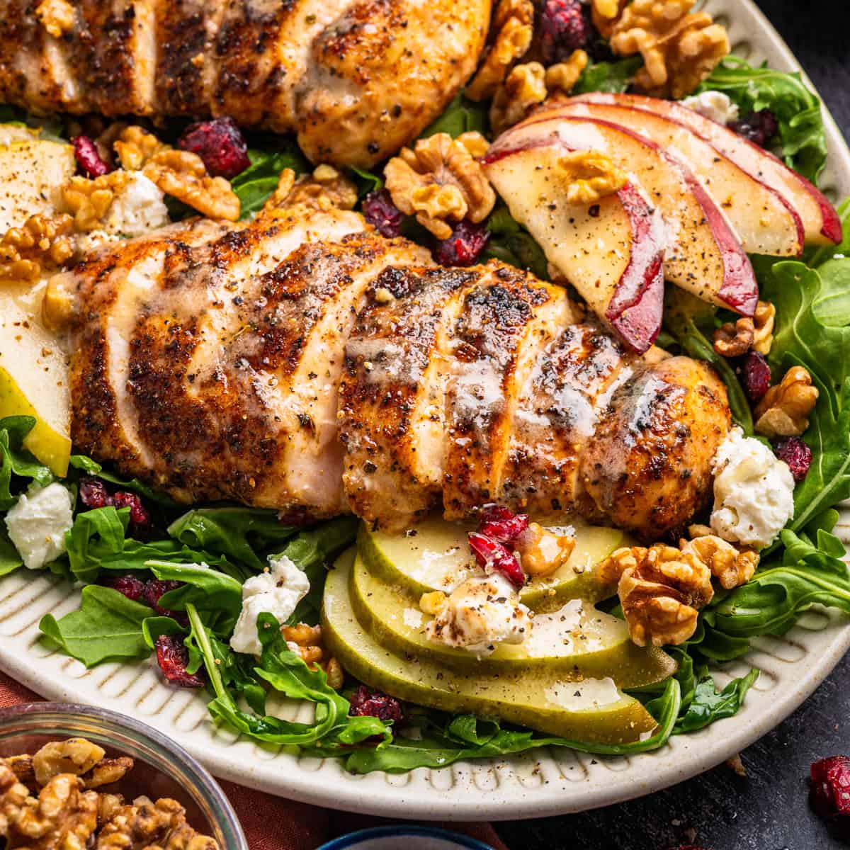 Chicken Arugula Salad Grilled chicken breast sliced on a bed of arugula with walnuts, sliced apples, crumbled cheese, and dried cranberries, all on a white plate.