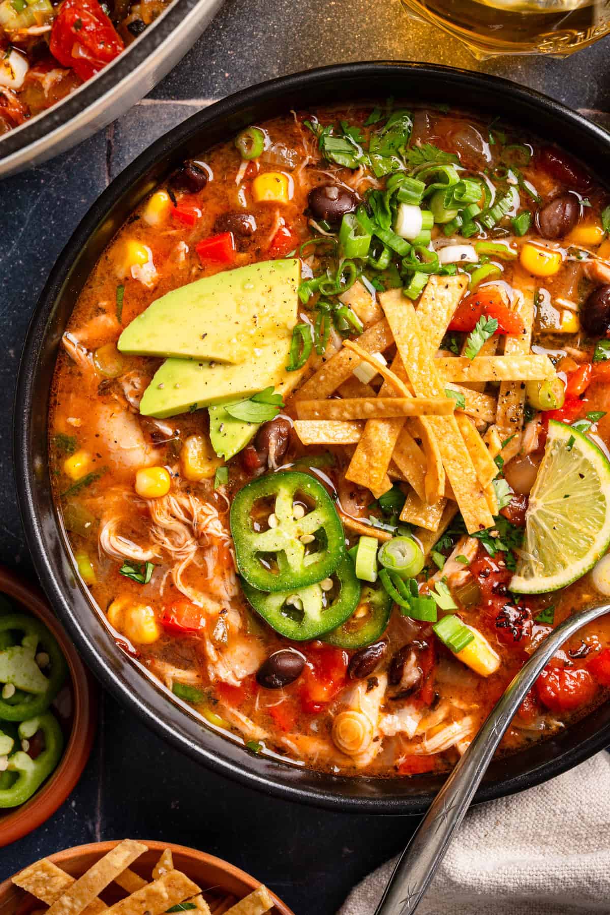 Chicken Tortilla Soup A bowl of chicken tortilla soup topped with avocado slices, jalapeño, crispy tortilla strips, lime wedge, scallions, , corn, tomatoes, and a spoon on the side.