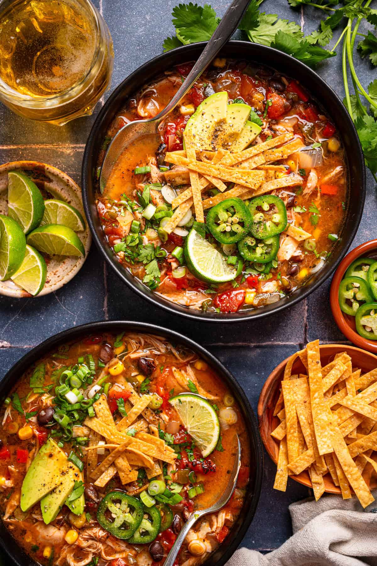 Chicken Tortilla Soup Two bowls of chicken tortilla soup topped with avocado, jalapeño, tortilla strips, lime, and green onions.