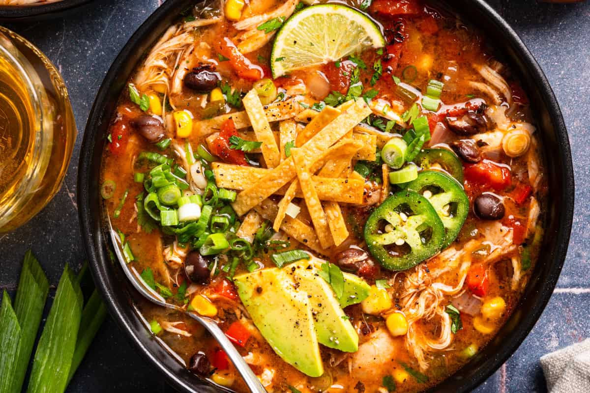 Chicken Tortilla Soup A bowl of chicken tortilla soup topped with sliced avocado, jalapeños, tortilla strips, green onions, corn, tomatoes, and a lime wedge.