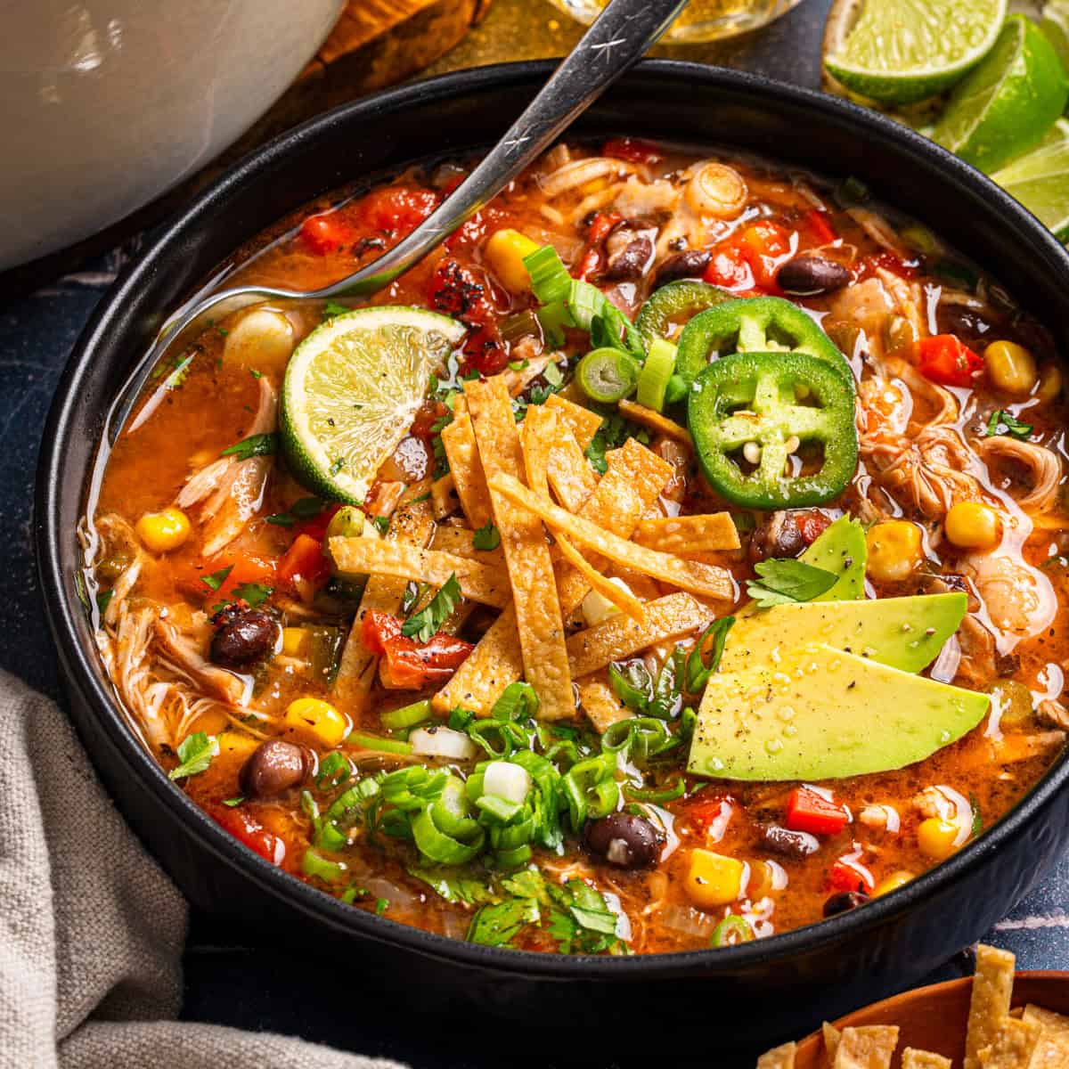 Chicken Tortilla Soup A bowl of tortilla soup topped with avocado slices, lime, jalapeño, tortilla strips, corn, chopped green onions, and fresh herbs.