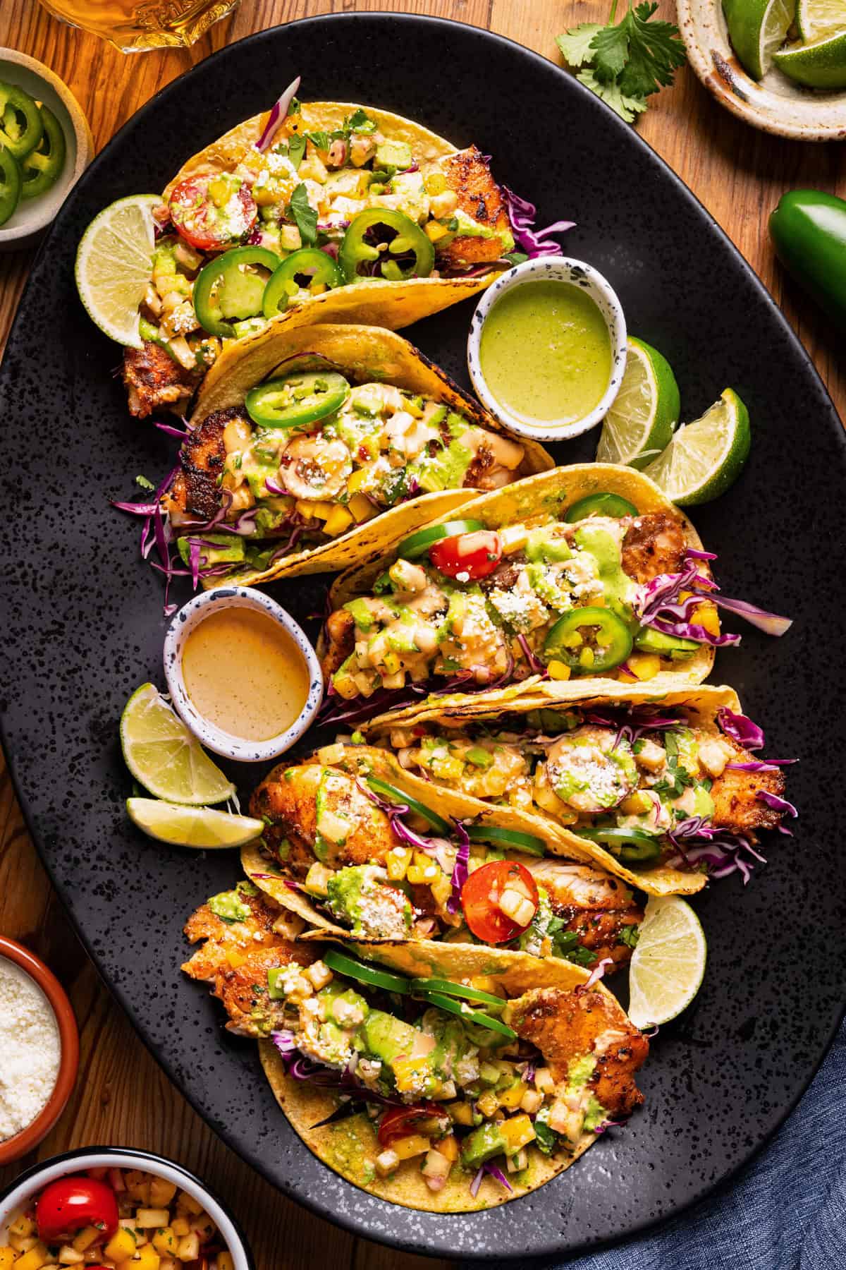 Oval black plate with six tacos filled with grilled fish, cabbage, salsa, jalape&ntilde;os, and avocado, garnished with lime wedges. 