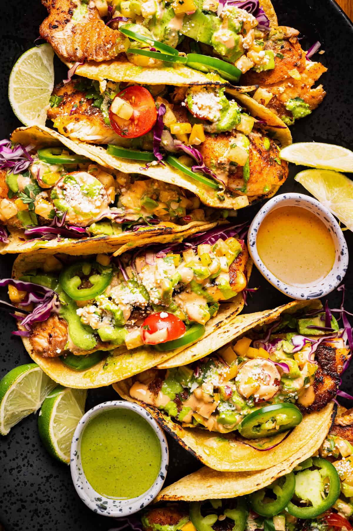 A close-up of grilled fish tacos in corn tortillas, topped with sliced jalape&ntilde;os, tomatoes, shredded cabbage, salsa, and drizzled with creamy sauces.