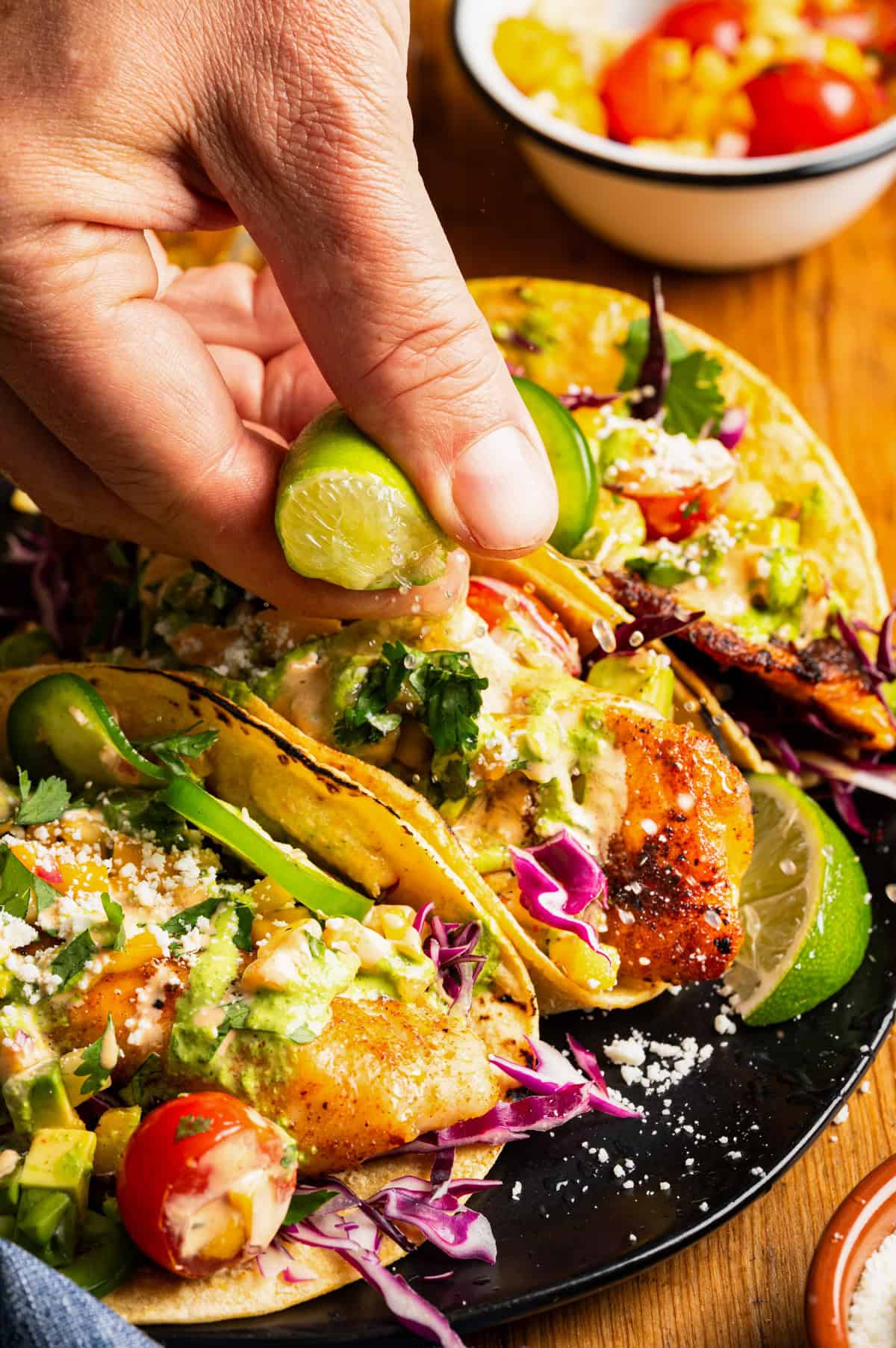 A hand squeezes a lime over tacos filled with fish, cabbage, tomatoes, jalape&ntilde;os, cilantro, and creamy sauce on a black plate.