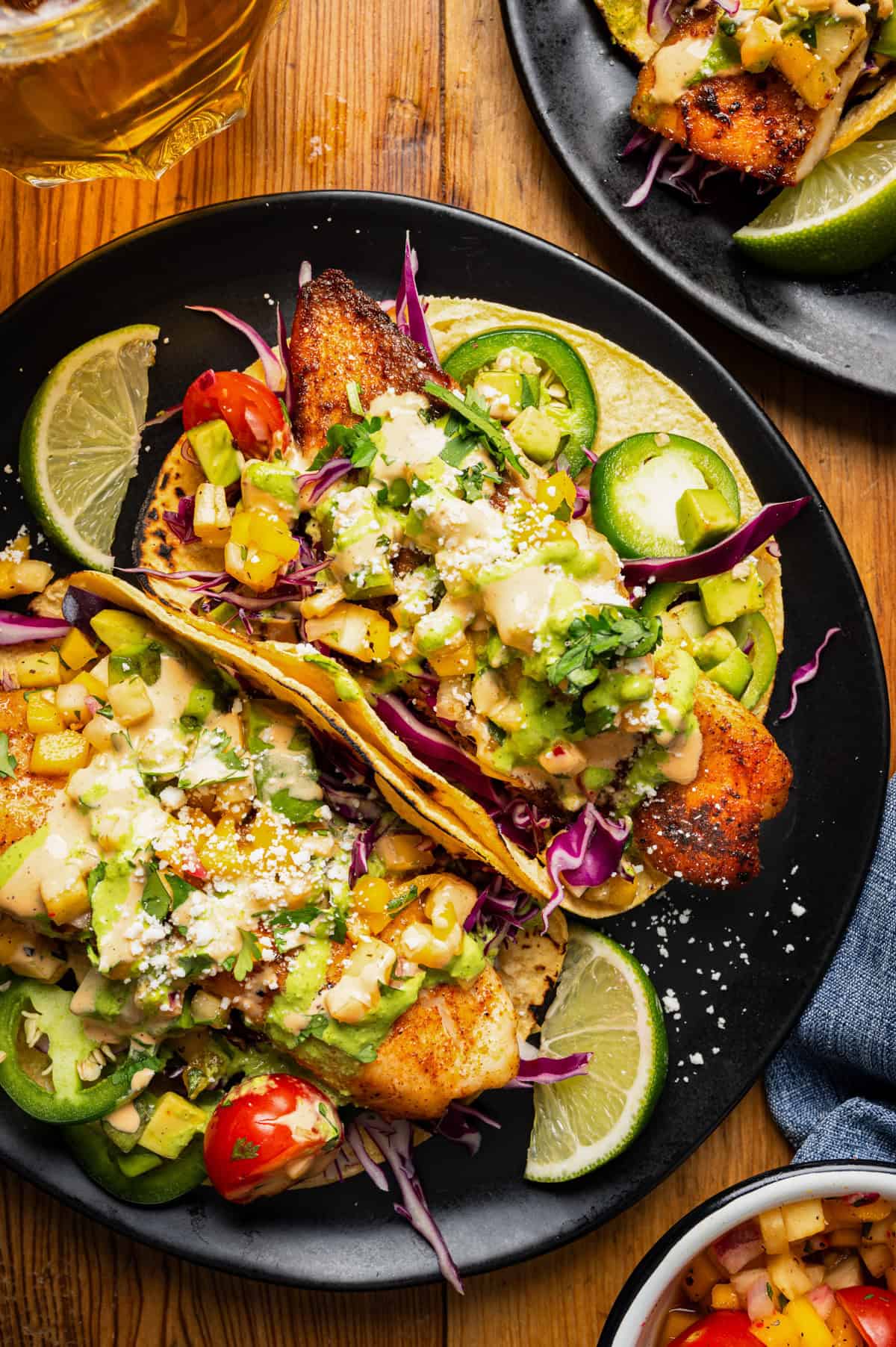 Fish tacos on a black plate with lime slices, cabbage, avocado, salsa, jalape&ntilde;o slices, and crumbled cheese, garnished with cilantro. 