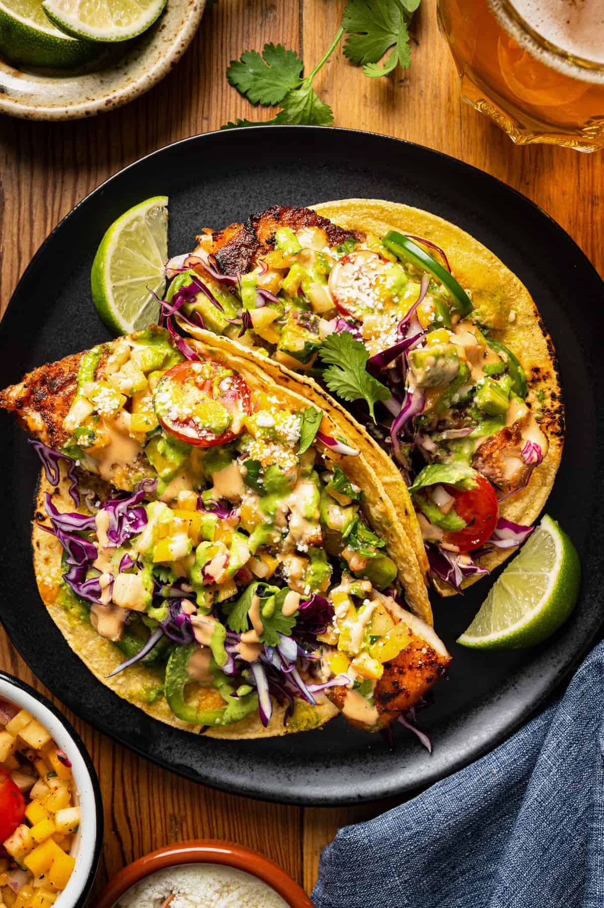 Two fish tacos on corn tortillas, topped with slaw, avocado, tomato, cilantro, sauce, and lime wedges, served on a black plate with a drink and salsa nearby on a wooden table.