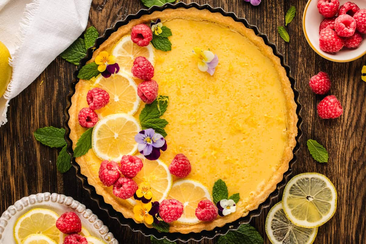 Lemon Tart A lemon tart topped with fresh raspberries, lemon slices, mint leaves, and edible flowers sits on a wooden table.