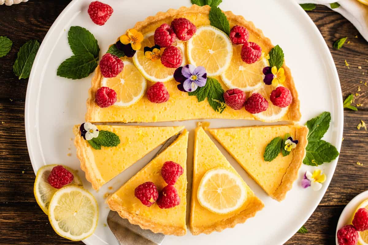 A lemon tart topped with fresh raspberries, lemon slices, mint leaves, and edible flowers sits on a round white plate. 