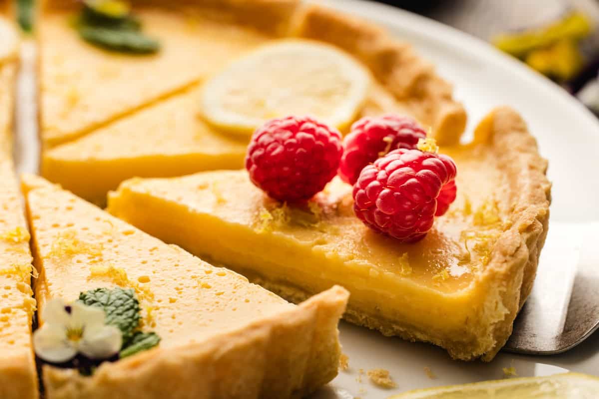 Lemon Tart A close-up of a lemon tart with a slice being served. The slice is topped with three raspberries and a lemon slice, and the tart is garnished with mint leaves and edible flowers.