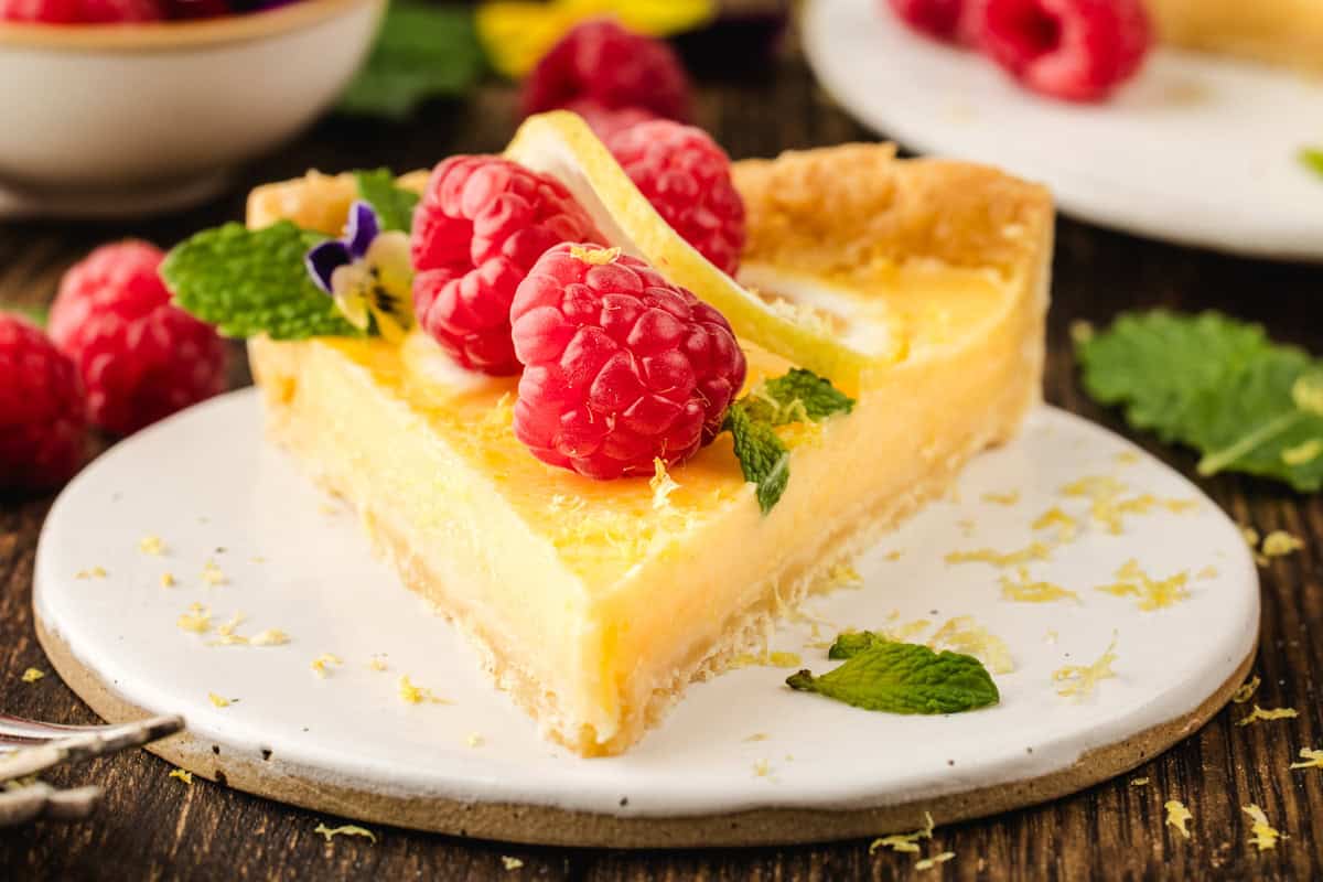 A slice of lemon tart topped with fresh raspberries, mint leaves, and a slice of lemon, served on a white plate.