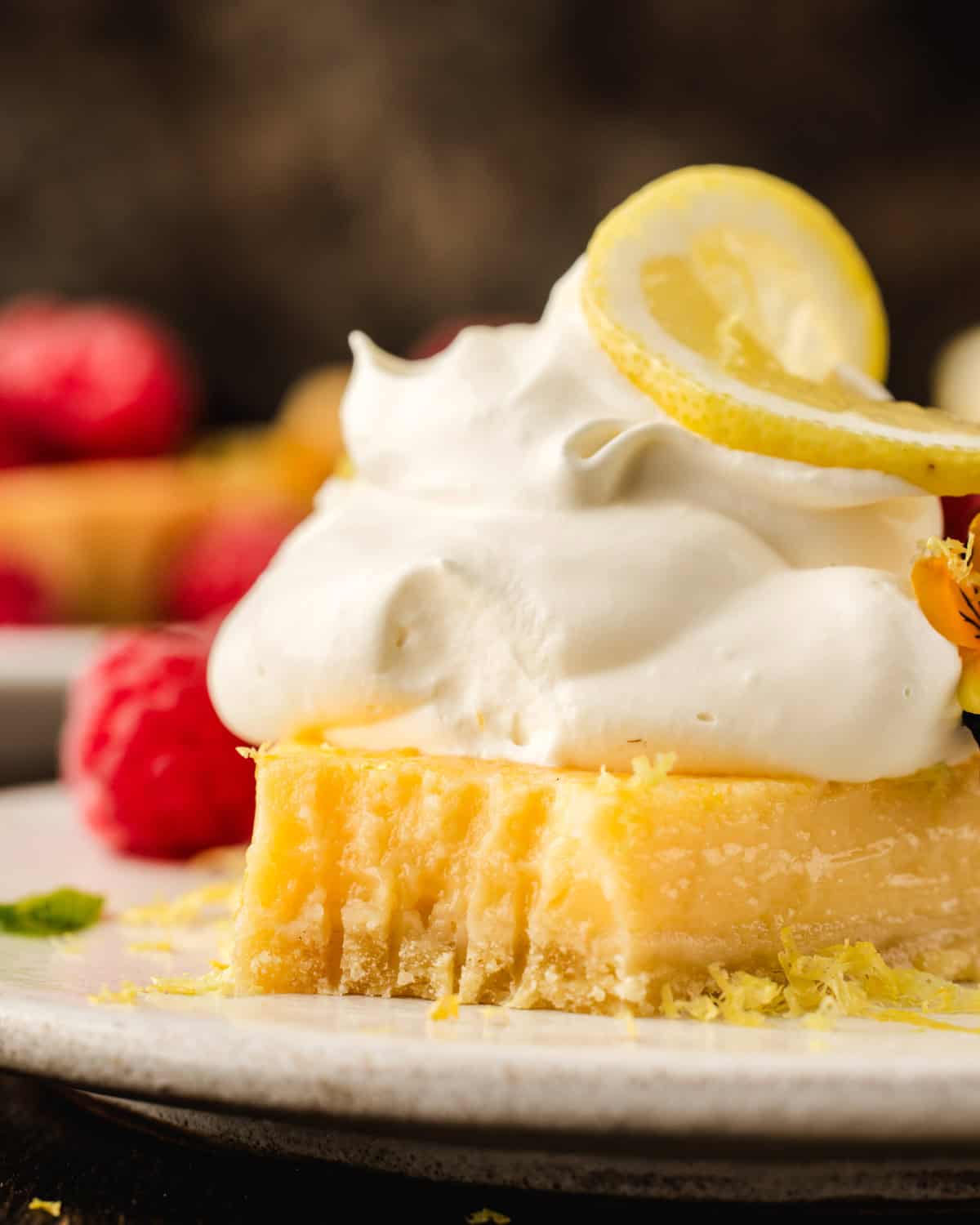 Lemon Tart A close-up of a lemon dessert topped with whipped cream and a lemon slice, with raspberries and lemon zest on a plate.