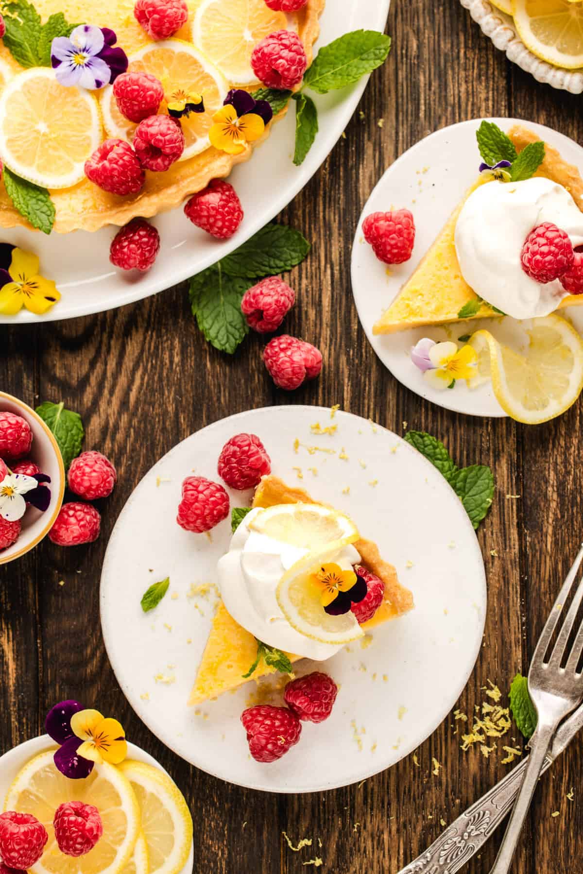 Lemon Tart Two plates with slices of lemon tart, topped with whipped cream, raspberries, edible flowers, and lemon slices.
