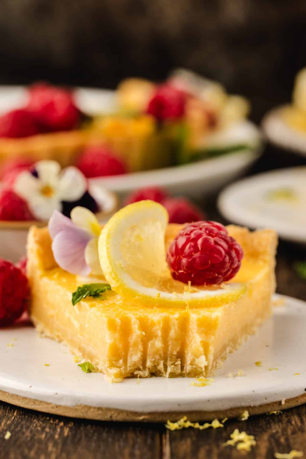 Lemon Tart A slice of lemon tart on a plate, topped with a lemon slice, a raspberry, edible flowers, and a mint leaf.