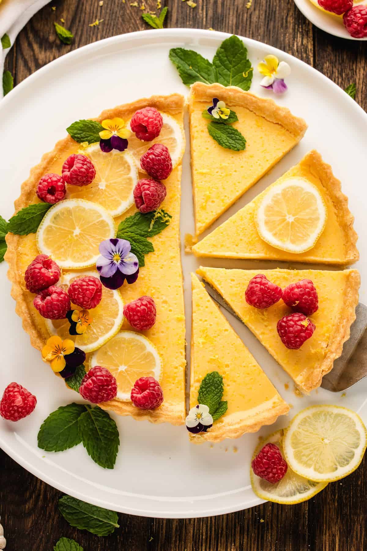 Lemon Tart A lemon tart on a white plate, garnished with fresh raspberries, lemon slices, mint leaves, and edible flowers.