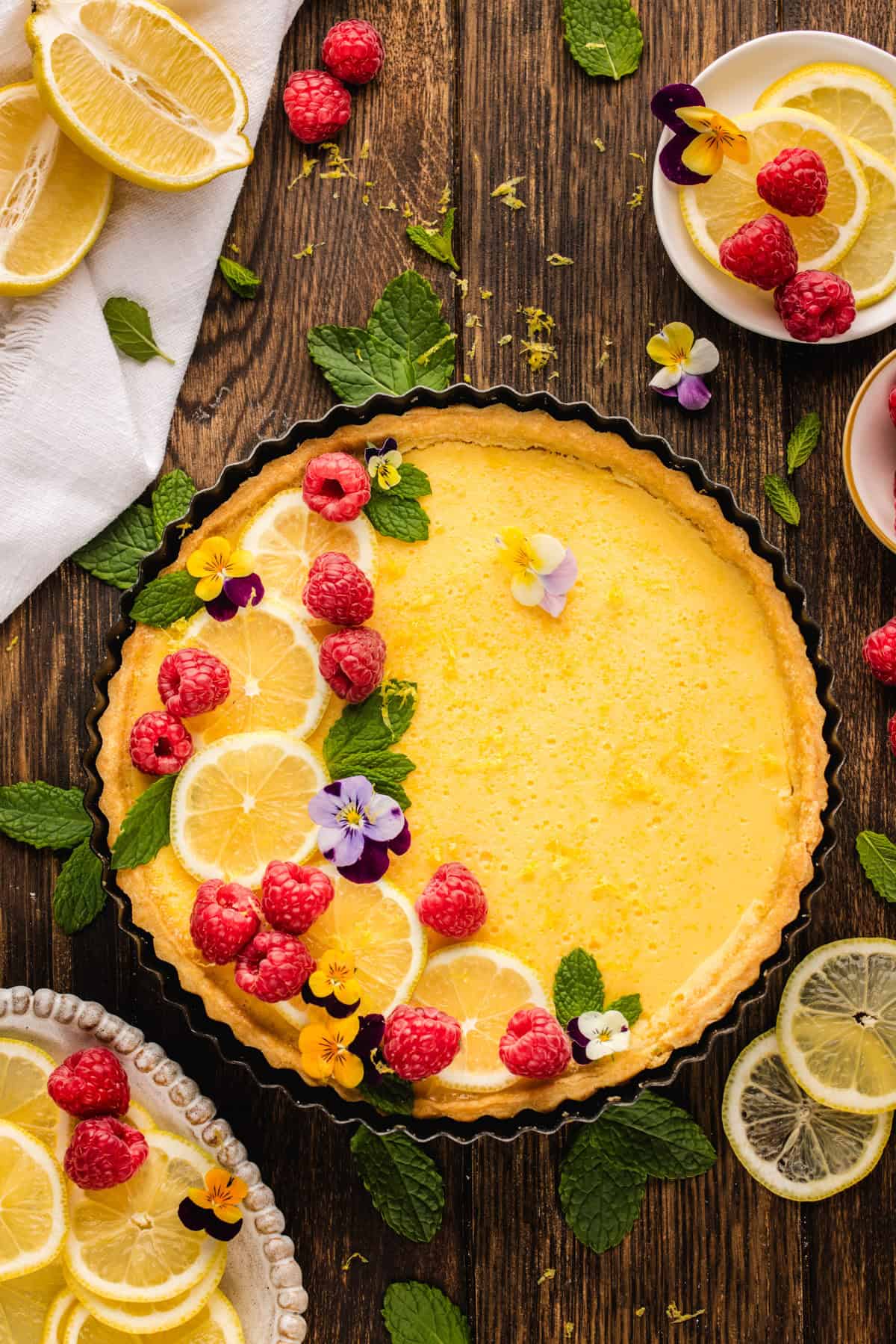 Lemon Tart A lemon tart decorated with fresh raspberries, lemon slices, mint leaves, and edible flowers sits on a wooden table.