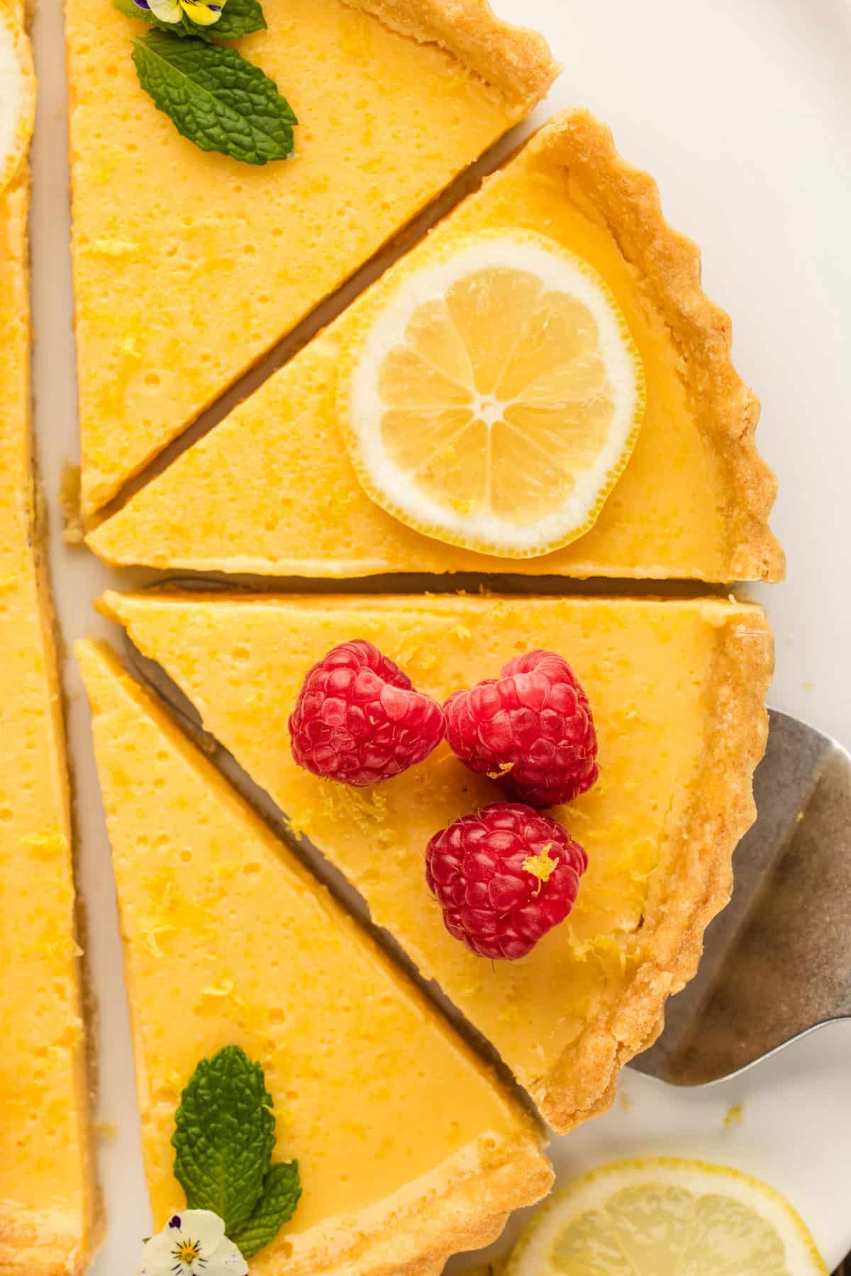 Lemon Tart A lemon tart sliced into pieces, with one slice topped with fresh raspberries and another with a lemon slice and mint leaves. The tart has a golden crust and creamy yellow filling.